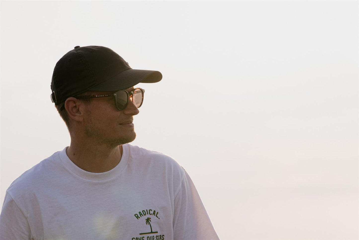 Man wearing a black cap and sunglasses with a white t-shirt featuring text and graphics against a light background