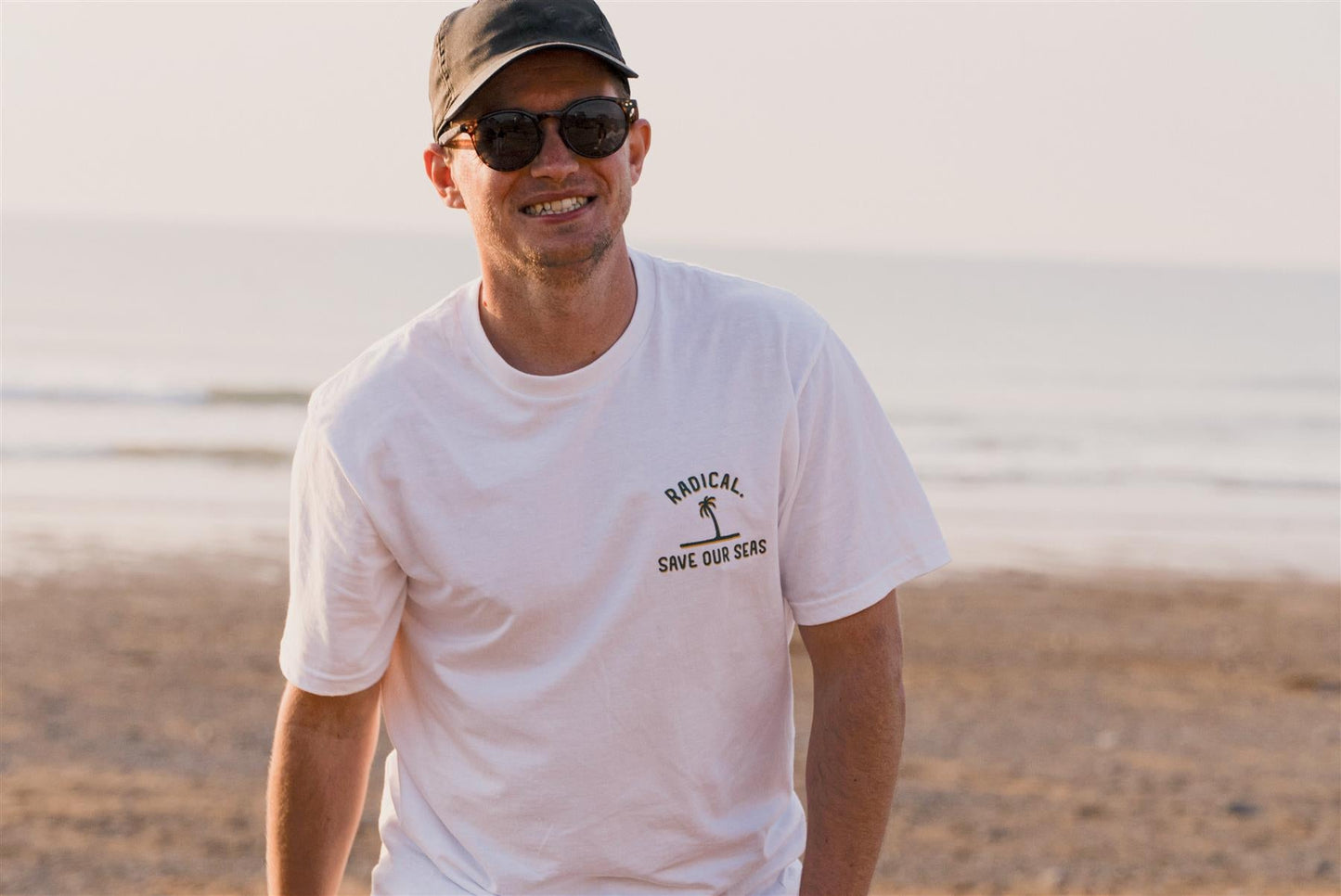Man wearing a white t-shirt with a logo and text, standing on a beach.
