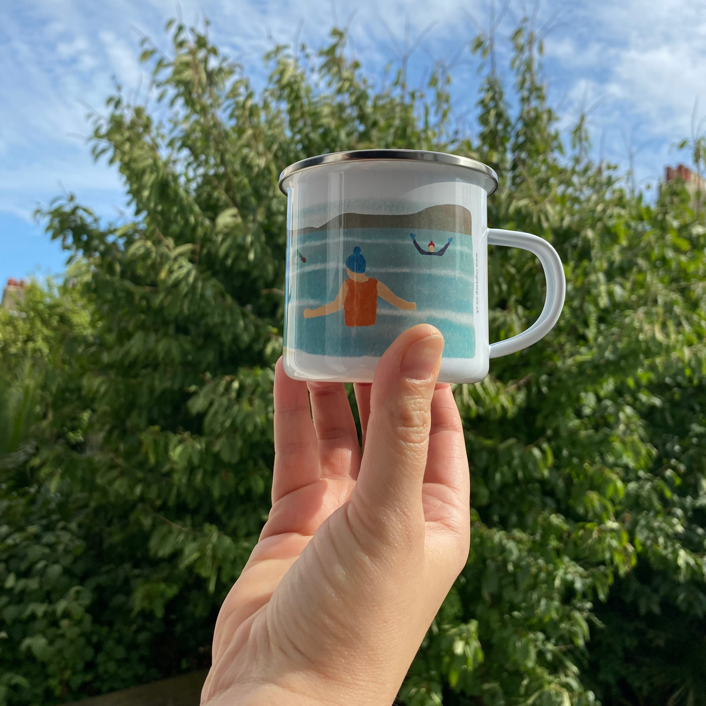 Hand holding a mug with a design of a person in a swimsuit against a blue sky, outdoors with trees in the background.