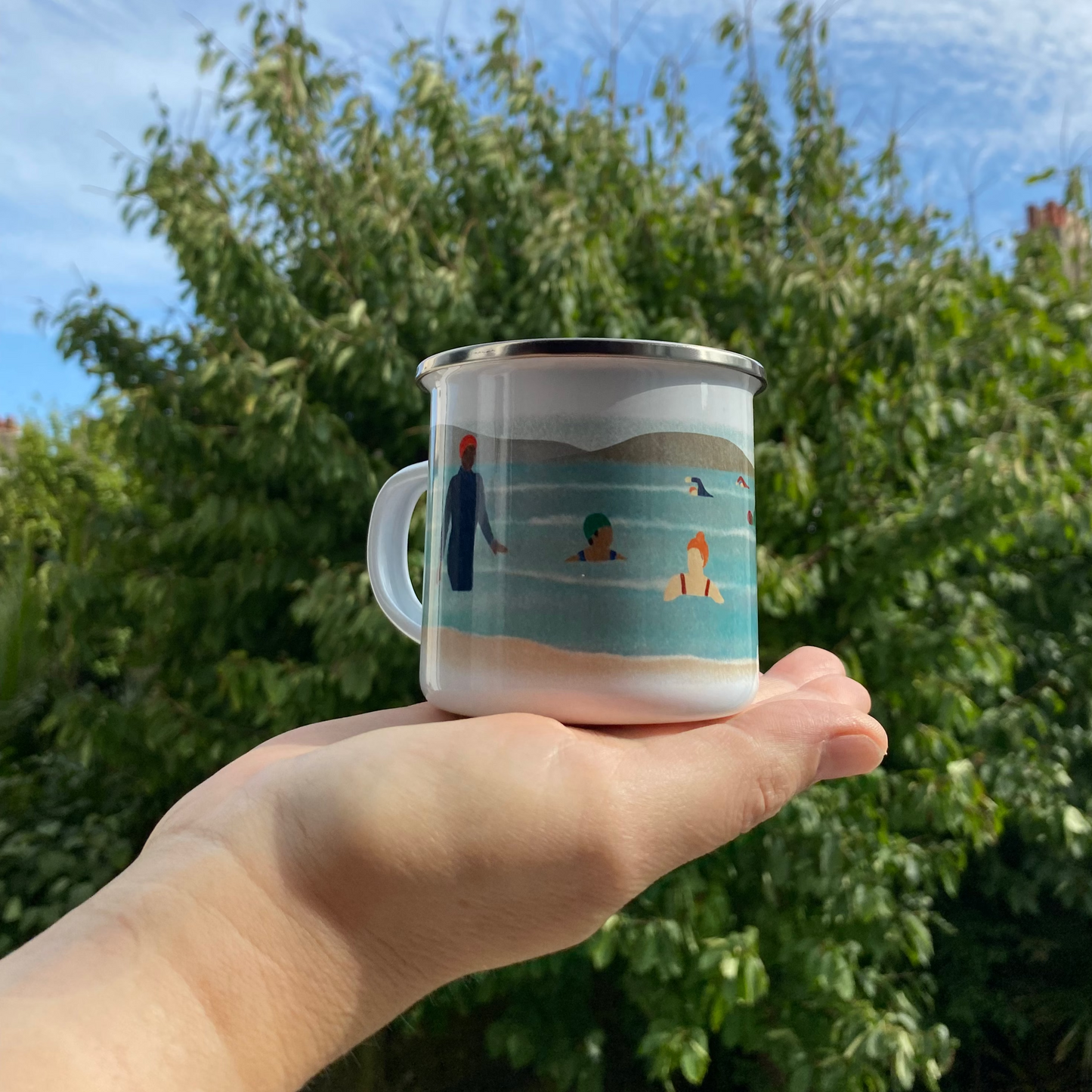 Hand holding a mug with a beach scene design against a green tree and blue sky background