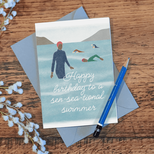 Birthday card with illustration of people swimming and text 'Happy birthday to a sea-sea- tional swimmer' on a wooden surface with a pencil and flowers.