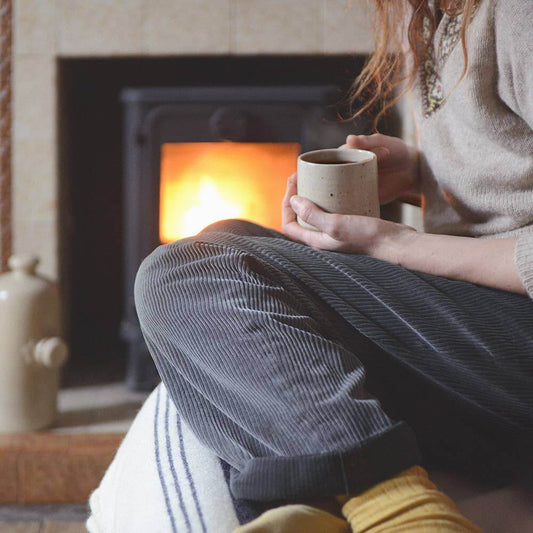 Person sitting by a fireplace holding a mug, wearing a sweater and socks.