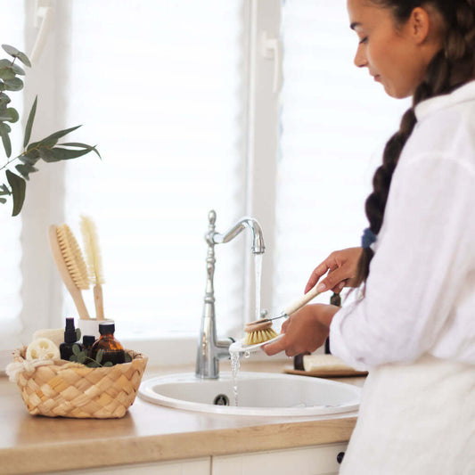 Woman washing a brush in a sink with a basket of cleaning supplies on a counter.