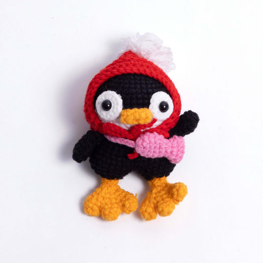 Crocheted penguin toy with a red hood and pink heart on a white background
