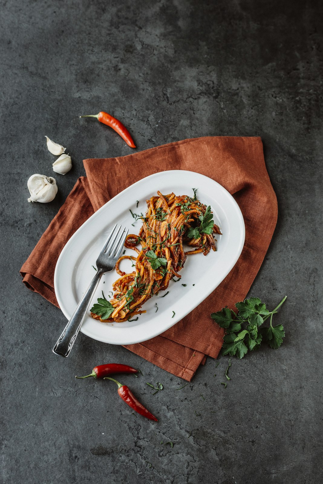 Plated dish of pasta with herbs on a dark surface with red peppers and garlic.