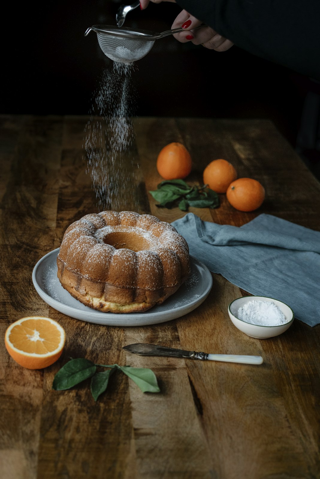 Bundt cake being dusted with powdered sugar on a wooden table with oranges and a knife.