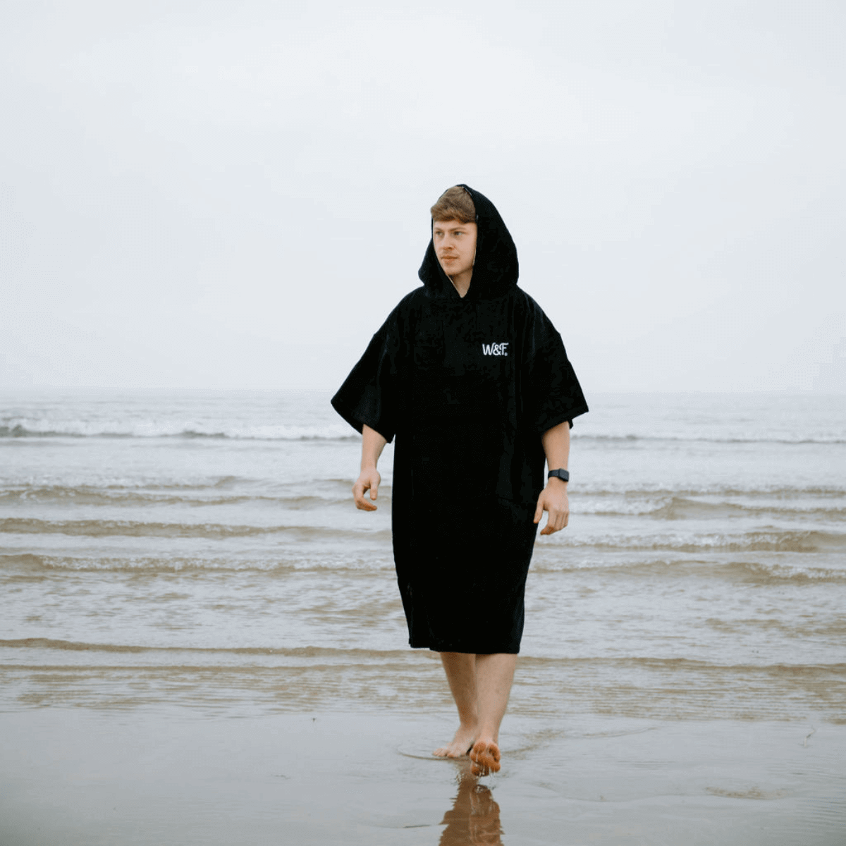 Man wearing a black hooded robe standing in open water from the Swim Wild collection.