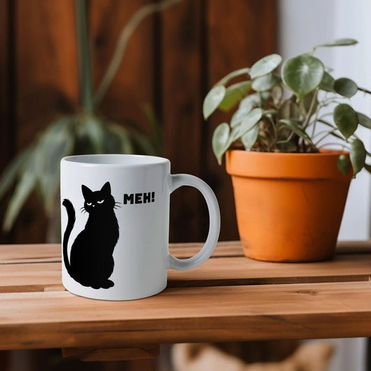 White mug with a black cat illustration and 'Meh!' text on a wooden table.