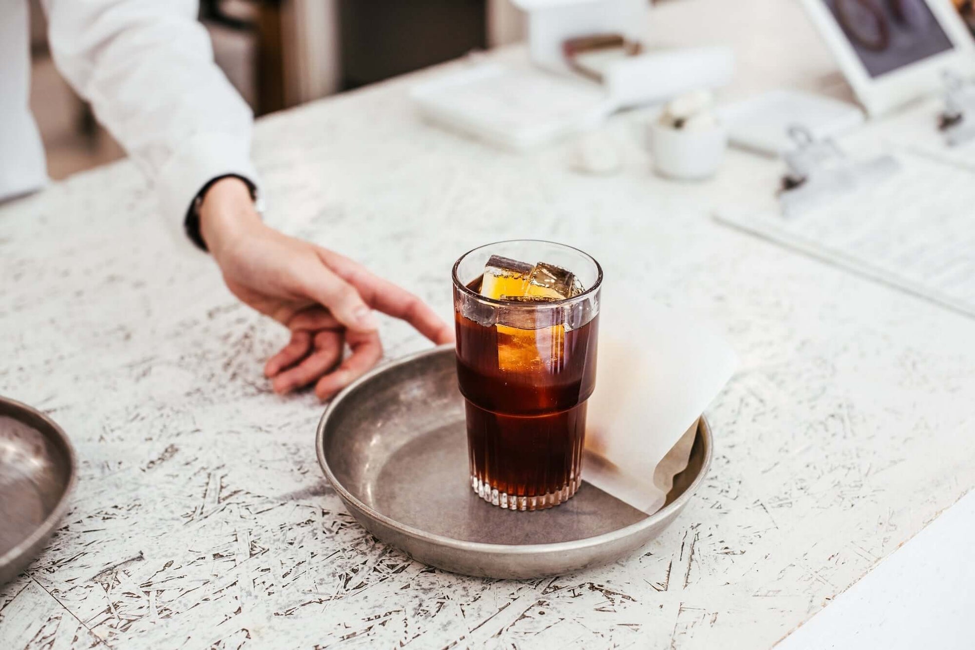 Glass of iced coffee on a tray with a hand reaching out, on a light-colored surface.