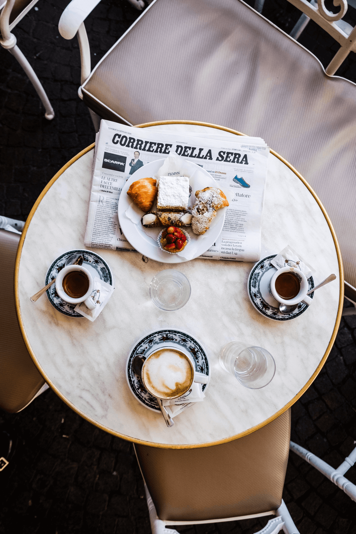 Round marble table with coffee cups, a newspaper, and pastries on a dark floor.