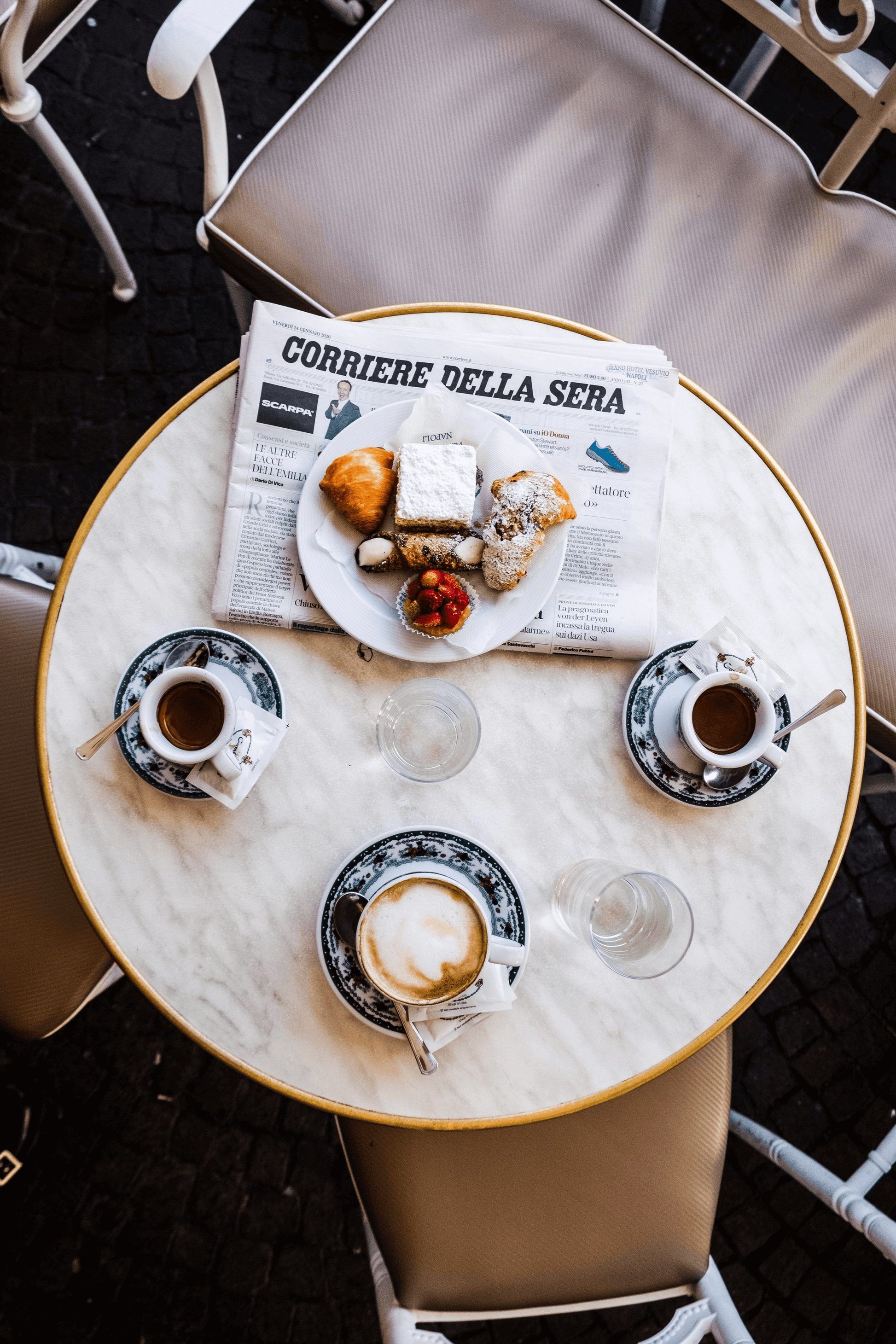 Round marble table with coffee cups, a newspaper, and pastries on a dark floor.