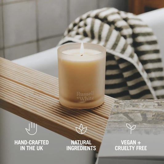 Candle on a wooden shelf with 'Russell + White' branding, surrounded by natural elements.