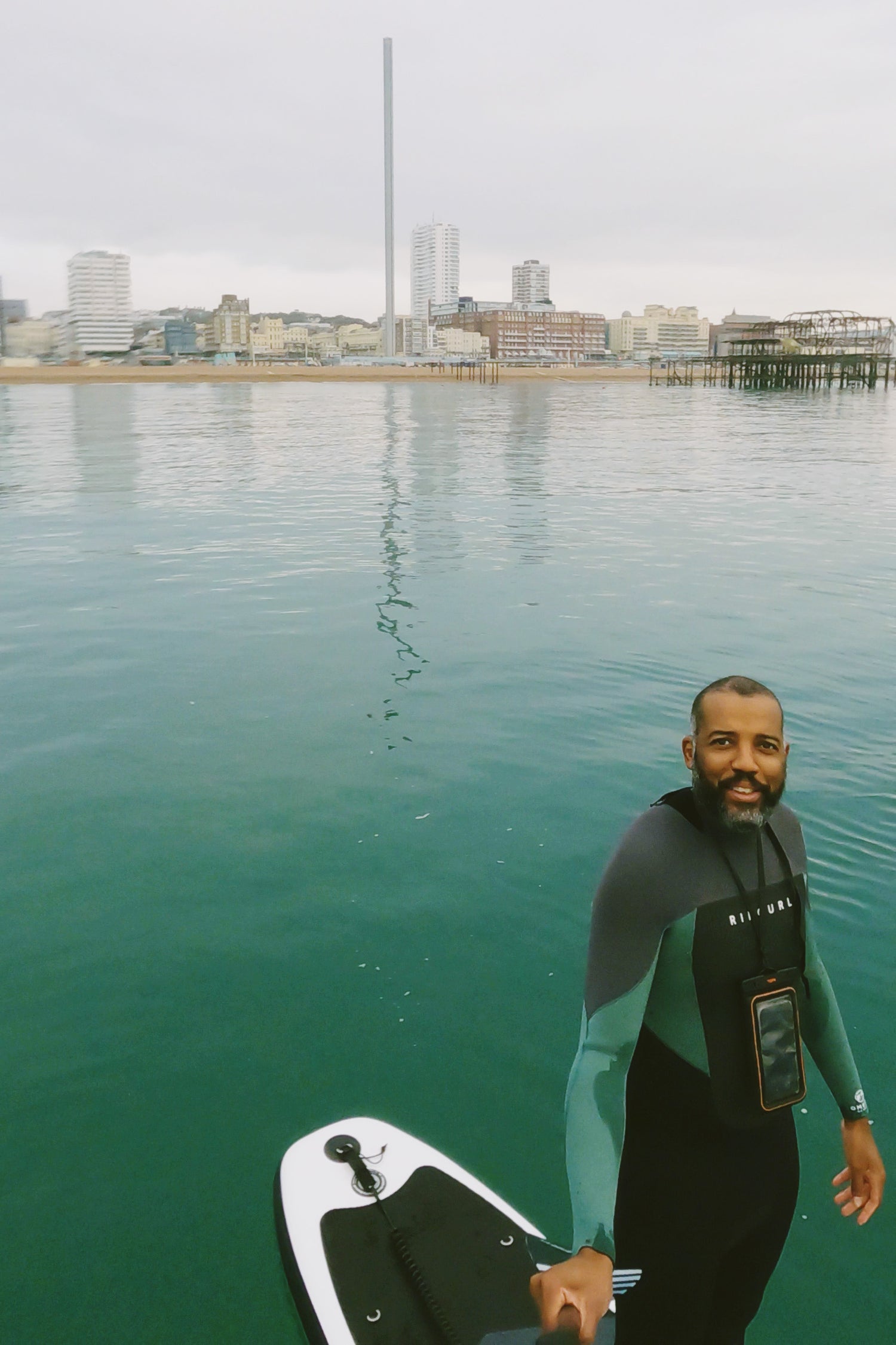 Man in a wetsuit holding a paddleboard on calm water with a cityscape in the background