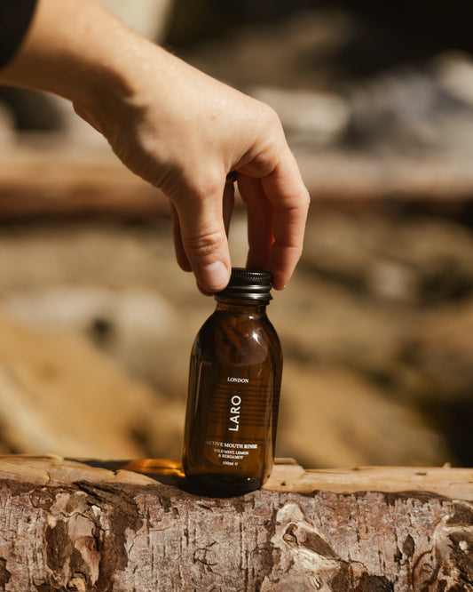 Hand holding a bottle of Travel Mouth Rinse, 100ml on a wooden surface outdoors.
