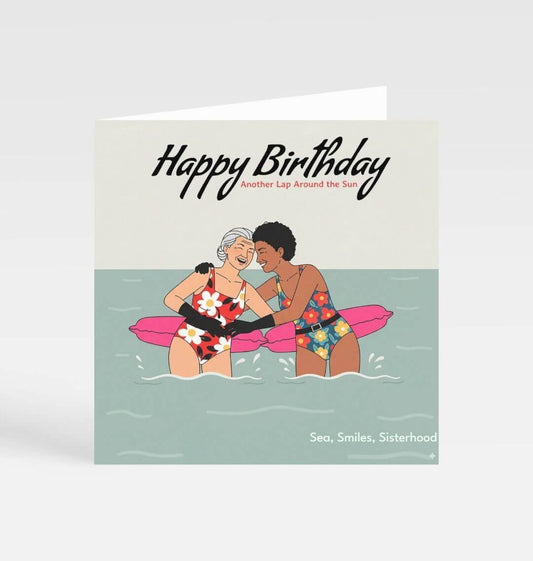 Birthday card with illustration of two women on a raft in the water, text 'Happy Birthday, Another Lap Around the Sun, Sea, Smiles, Sisterhood'.