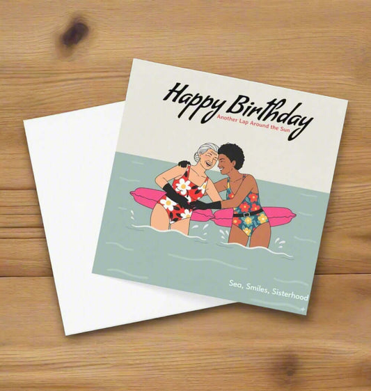 Birthday card with illustration of two people on a raft, text 'Happy Birthday, Another Lap Around the Sun', and 'Sea, Smiles, Sisterhood' on a light green background.