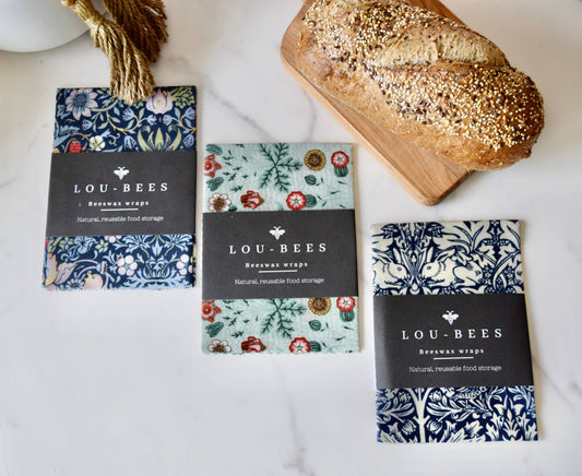 Three packages of Lou-Bees beeswax wraps on a white surface with a loaf of bread.
