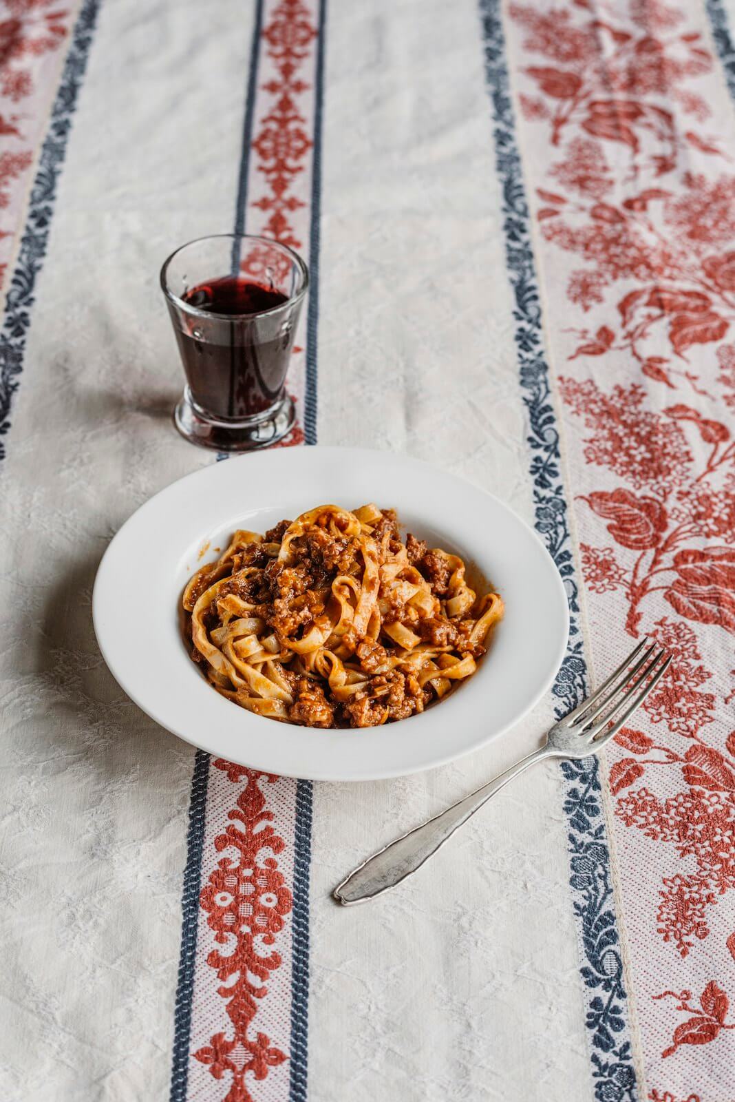 Delicious plate of pasta with sauce next to a glass of red wine on a patterned tablecloth, representing Originale: Recipes and Essentials of Italian Cooking.
