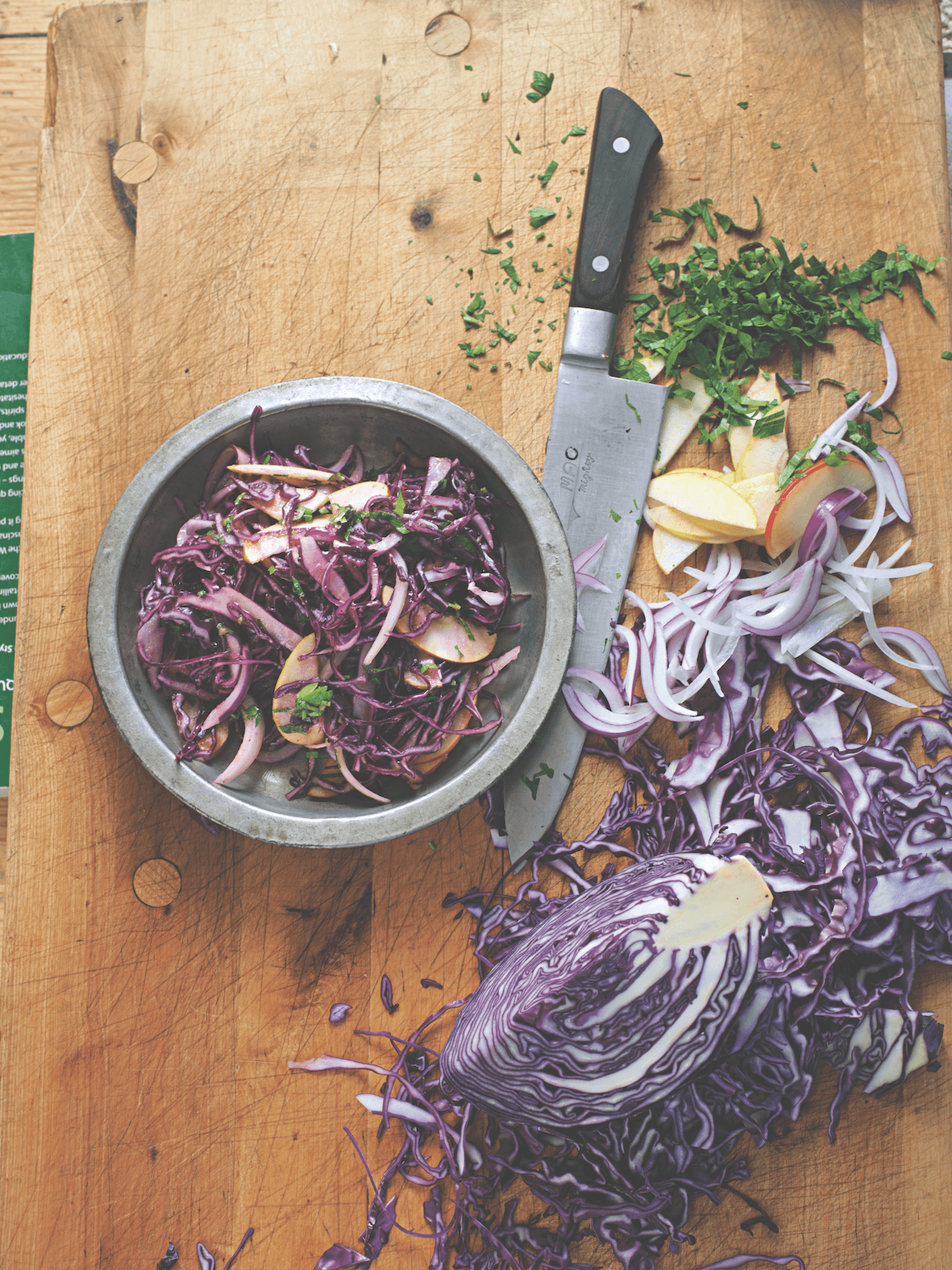 Sliced red cabbage on a wooden cutting board with a knife and bowl of salad.