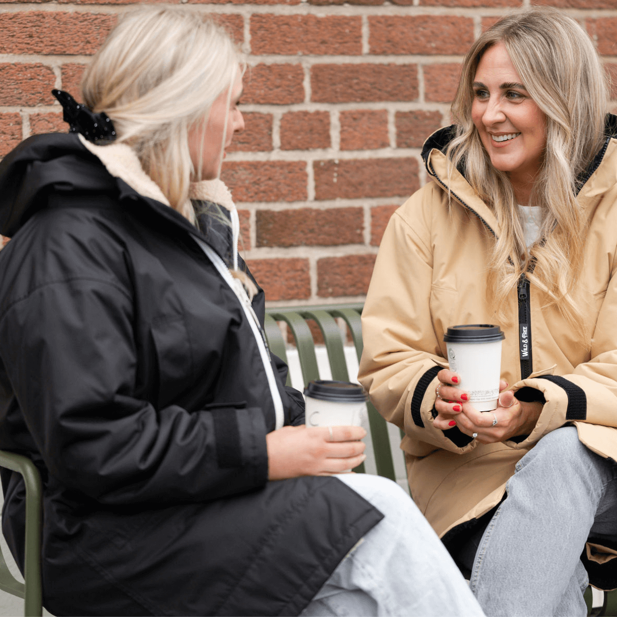 Women in stylish coats enjoying coffee, featuring the Raincoat & Changing Robe in One - The Classic Cobe Black.