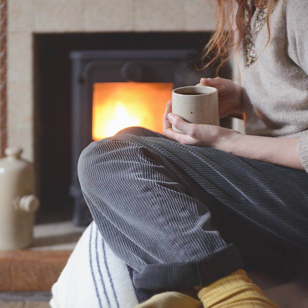 Person sitting by a fireplace holding a mug, wearing a sweater and socks.