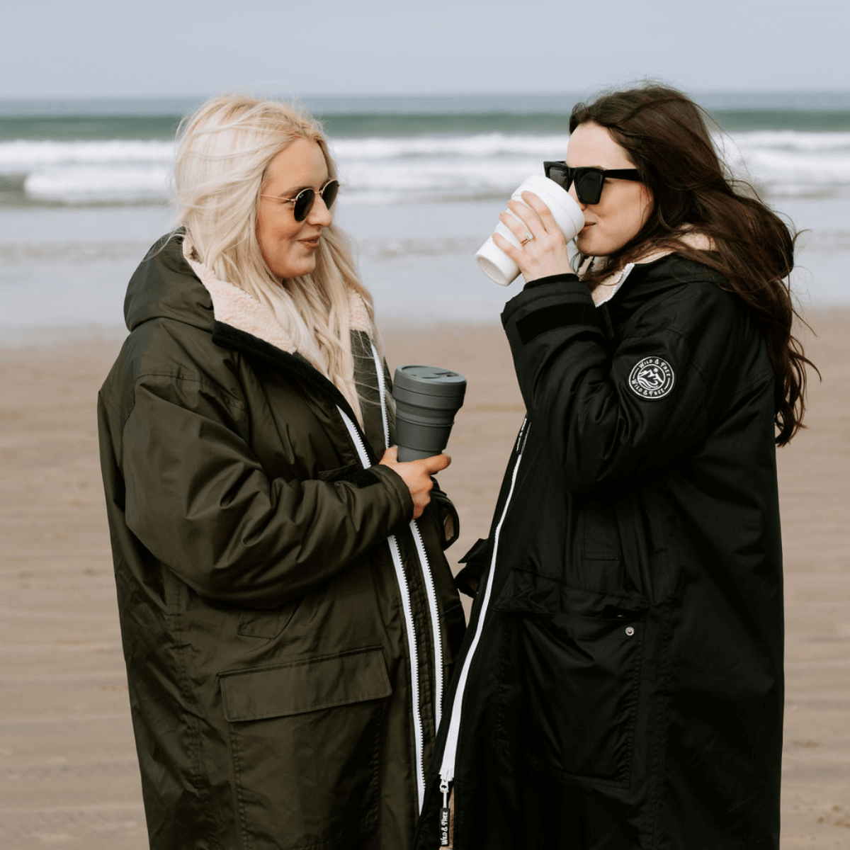 Two women wearing the Raincoat & Changing Robe in One - The Classic Cobe Black by the beach, enjoying drinks.