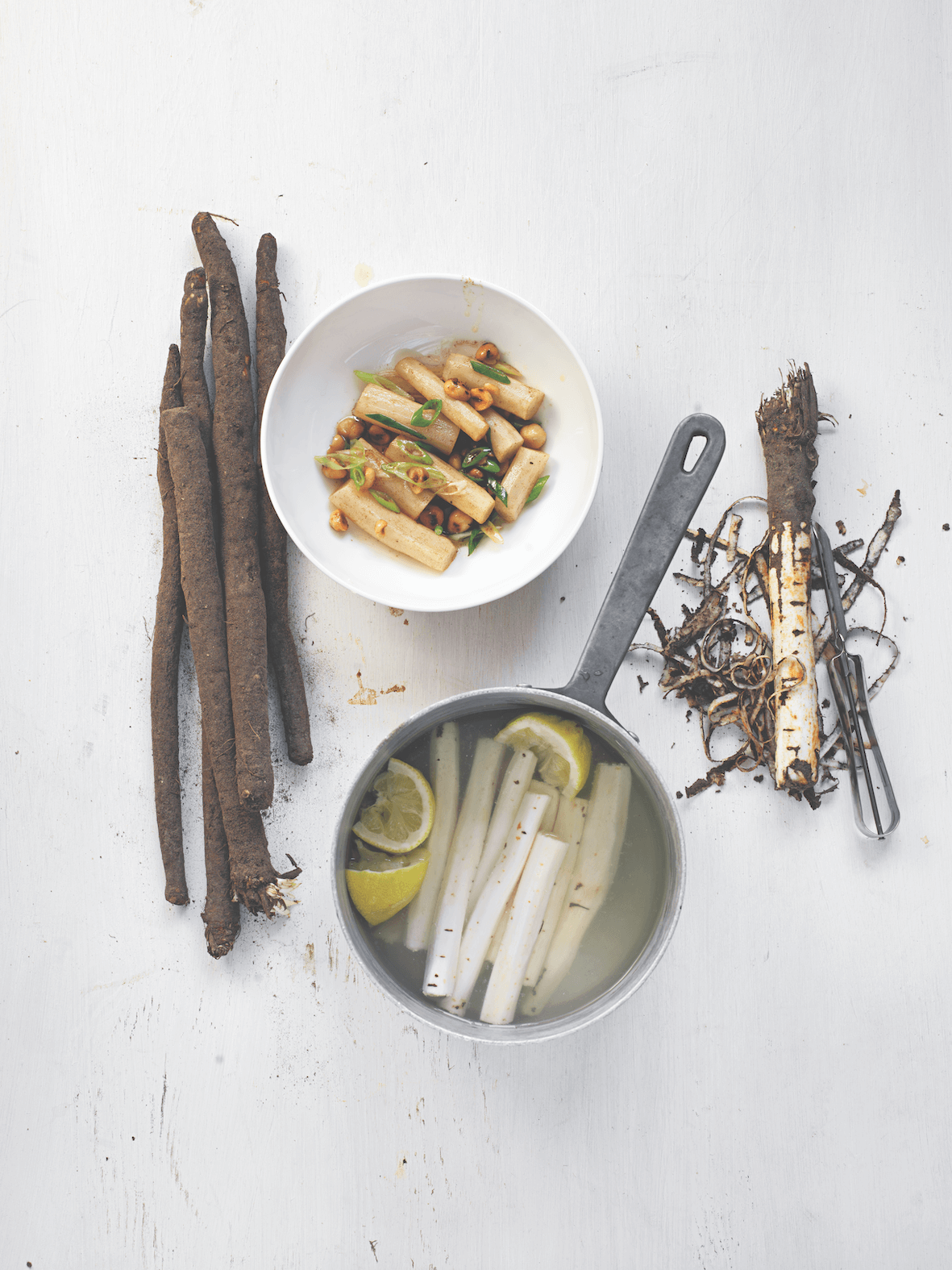 White bowl with food, pot with lemons and white sticks, and dried roots on a light surface