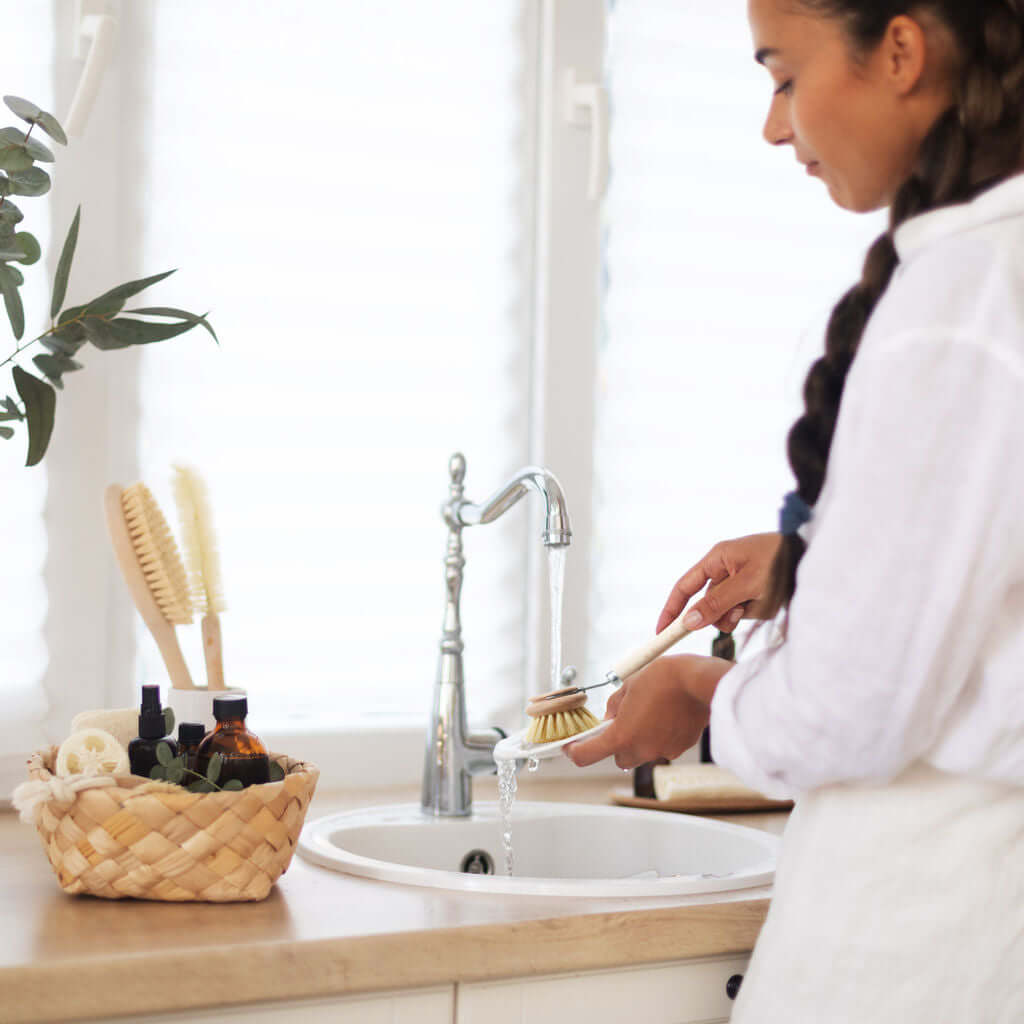 Woman washing a brush in a sink with a basket of cleaning supplies on a counter.