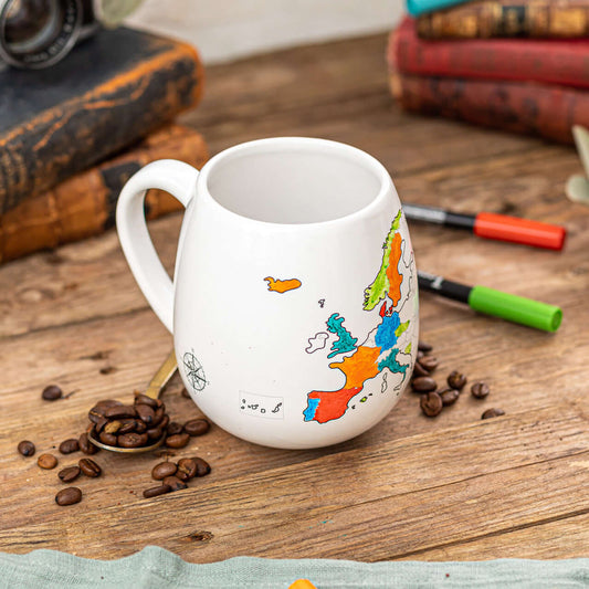 White mug with a colorful map design on a wooden table with coffee beans and books.
