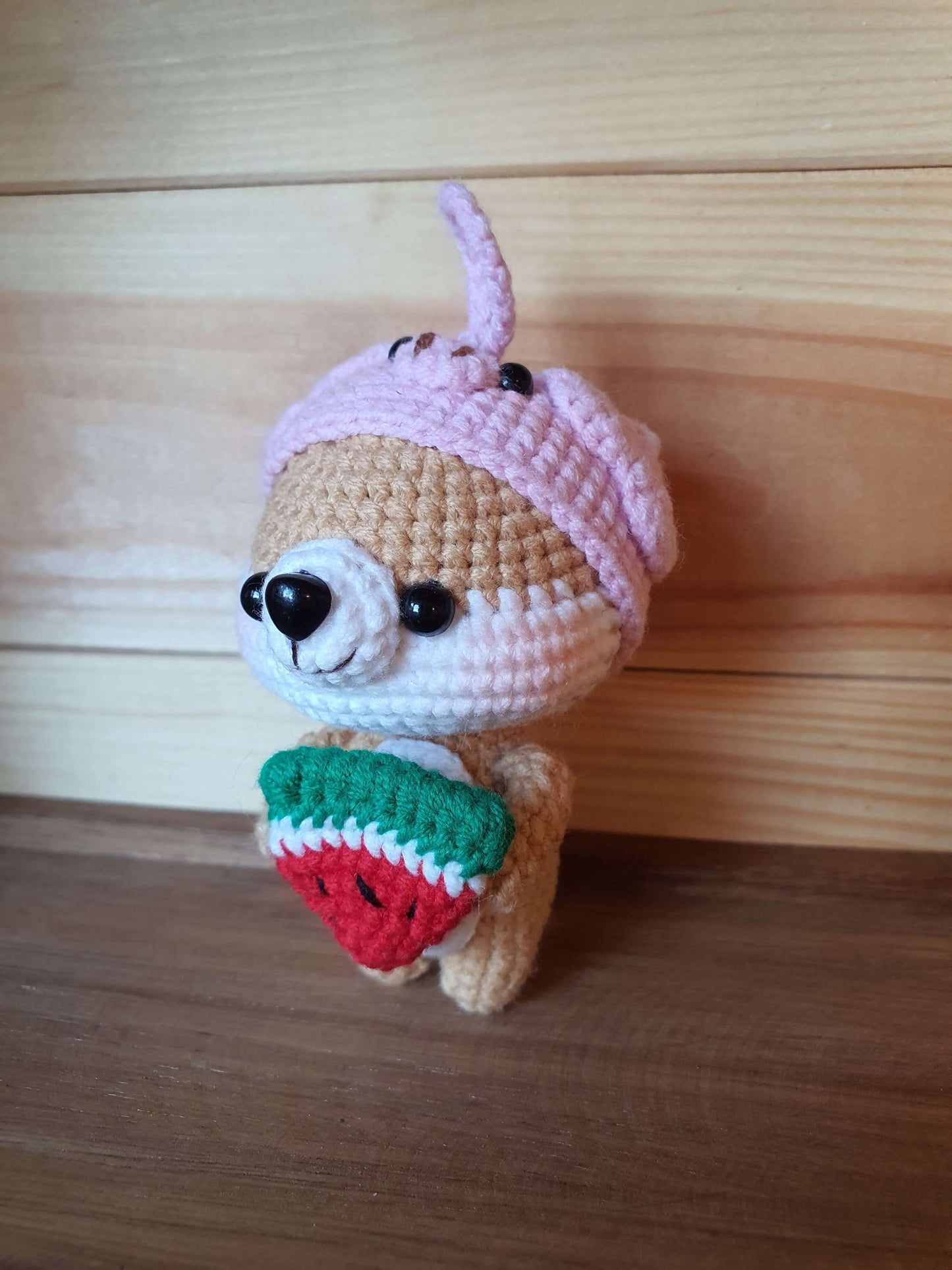 Crocheted animal toy with a pink hat and green and red body on a wooden surface