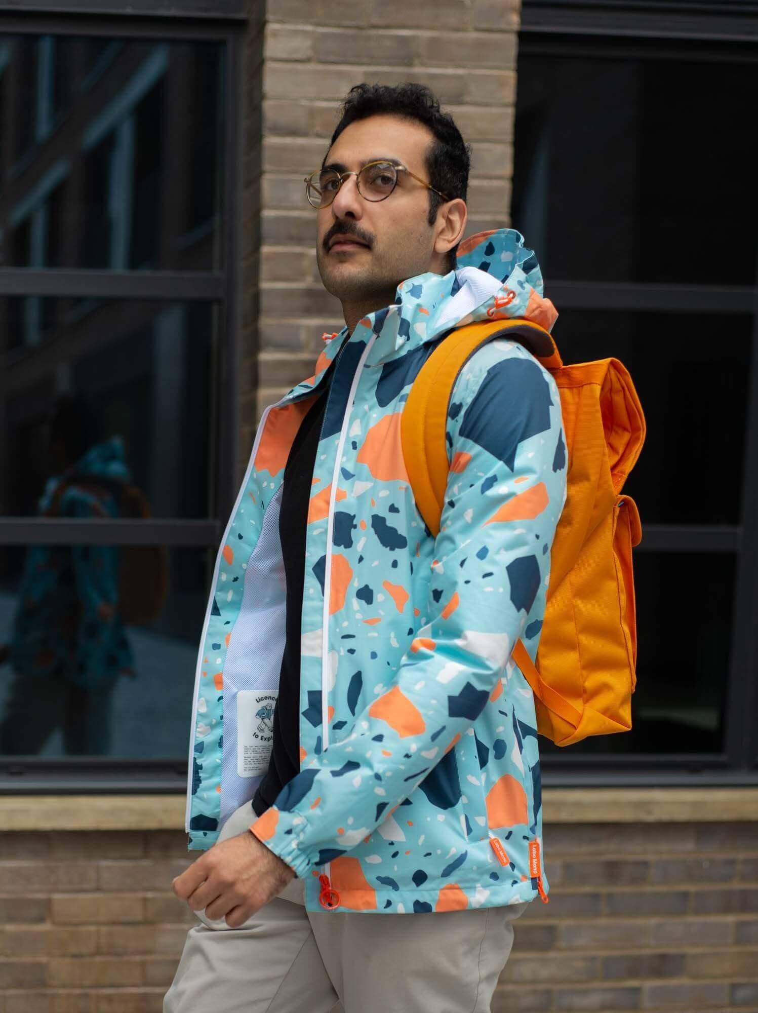 Man wearing a colorful printed jacket and orange backpack, walking confidently outdoors.