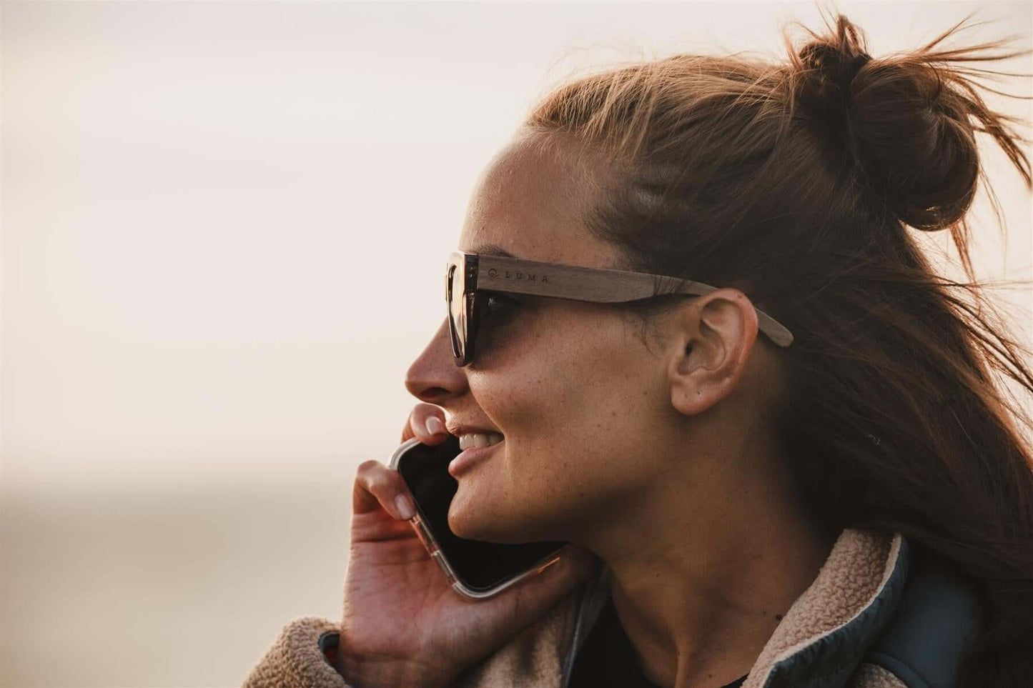 Luma Sunglasses - Polzeath - Smoke worn by a woman talking on the phone, enjoying coastal scenery.