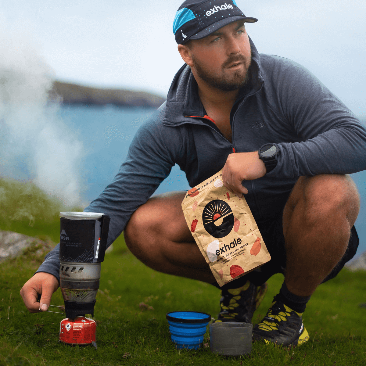 Man preparing Organic Dark(ish) Roast Coffee outdoors near a camping stove with beautiful scenery.