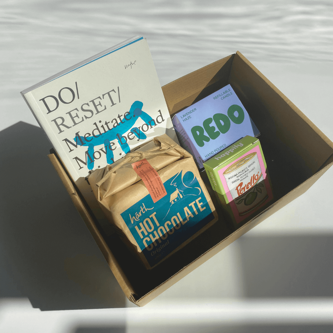 Open gift box containing a book, hot chocolate, REDO vegetable wax candle, and mini puzzle with visible branding on a light background.