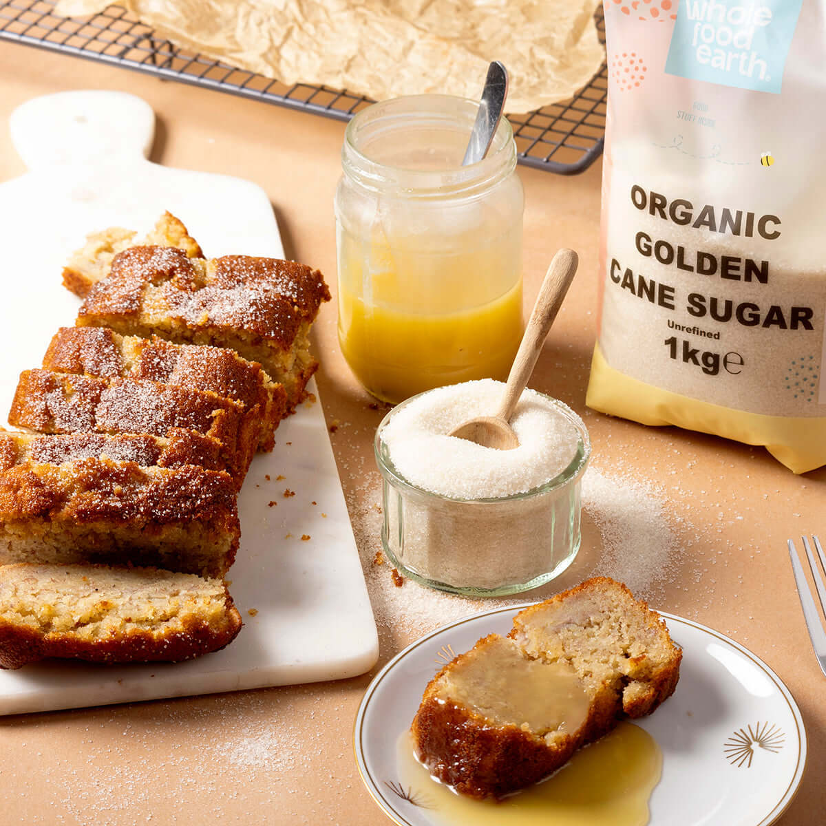 Loaf of bread with slices, jar of honey, and bag of organic golden cane sugar on a wooden surface.