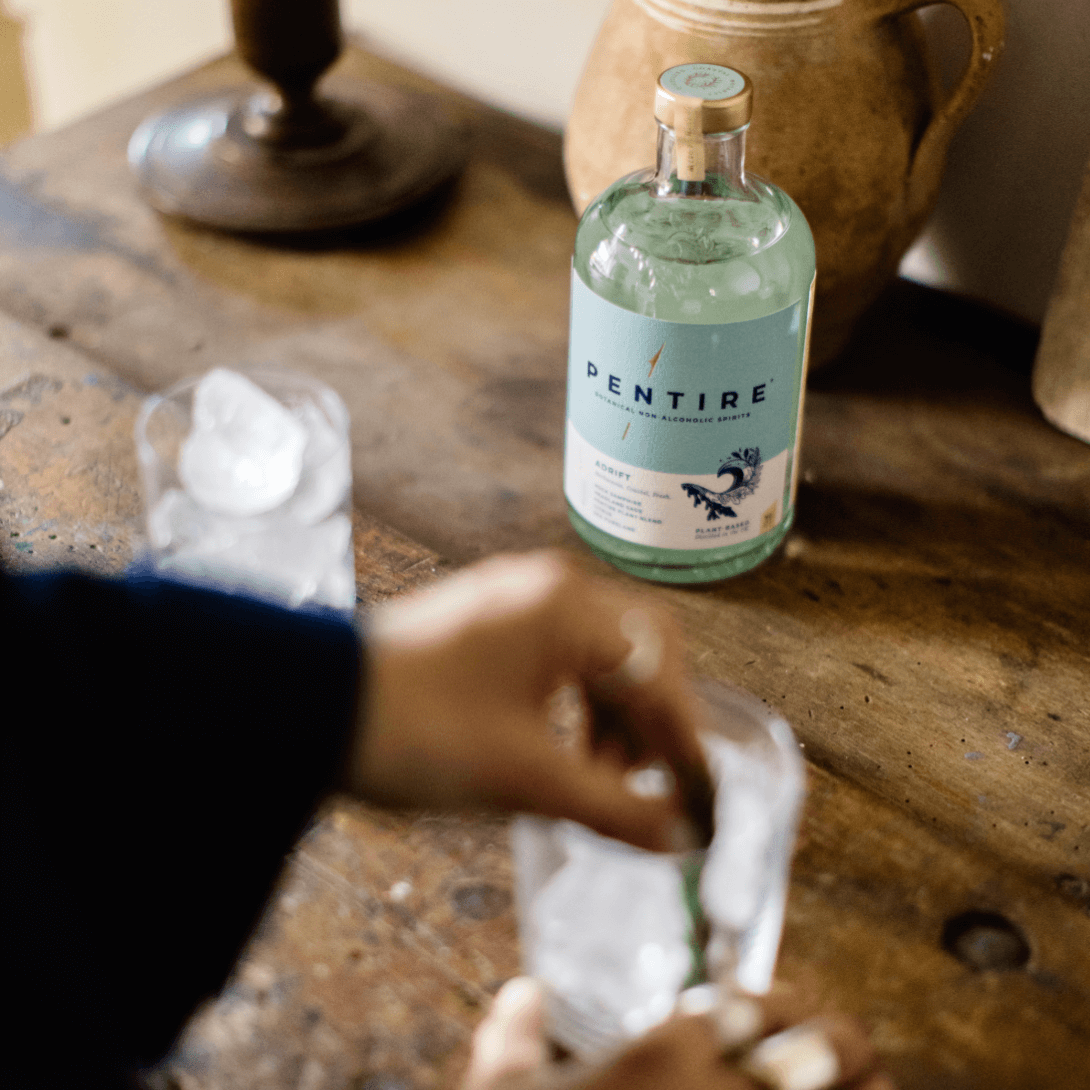 A hand preparing a drink with Pentire Adrift non-alcoholic spirit on a wooden table with ice.