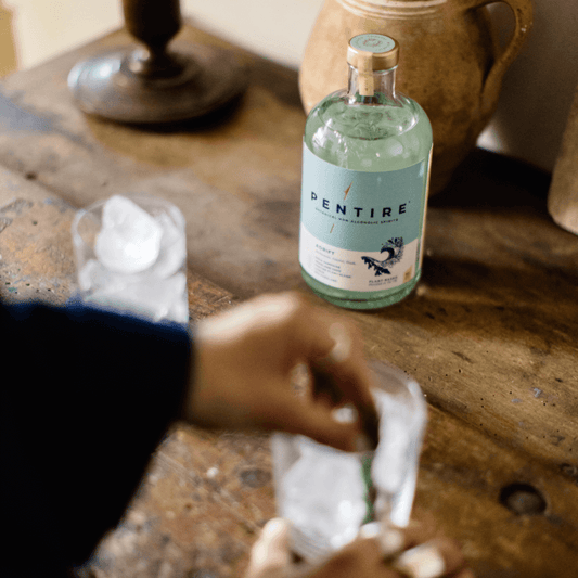 A hand preparing a drink with Pentire Adrift non-alcoholic spirit on a wooden table with ice.