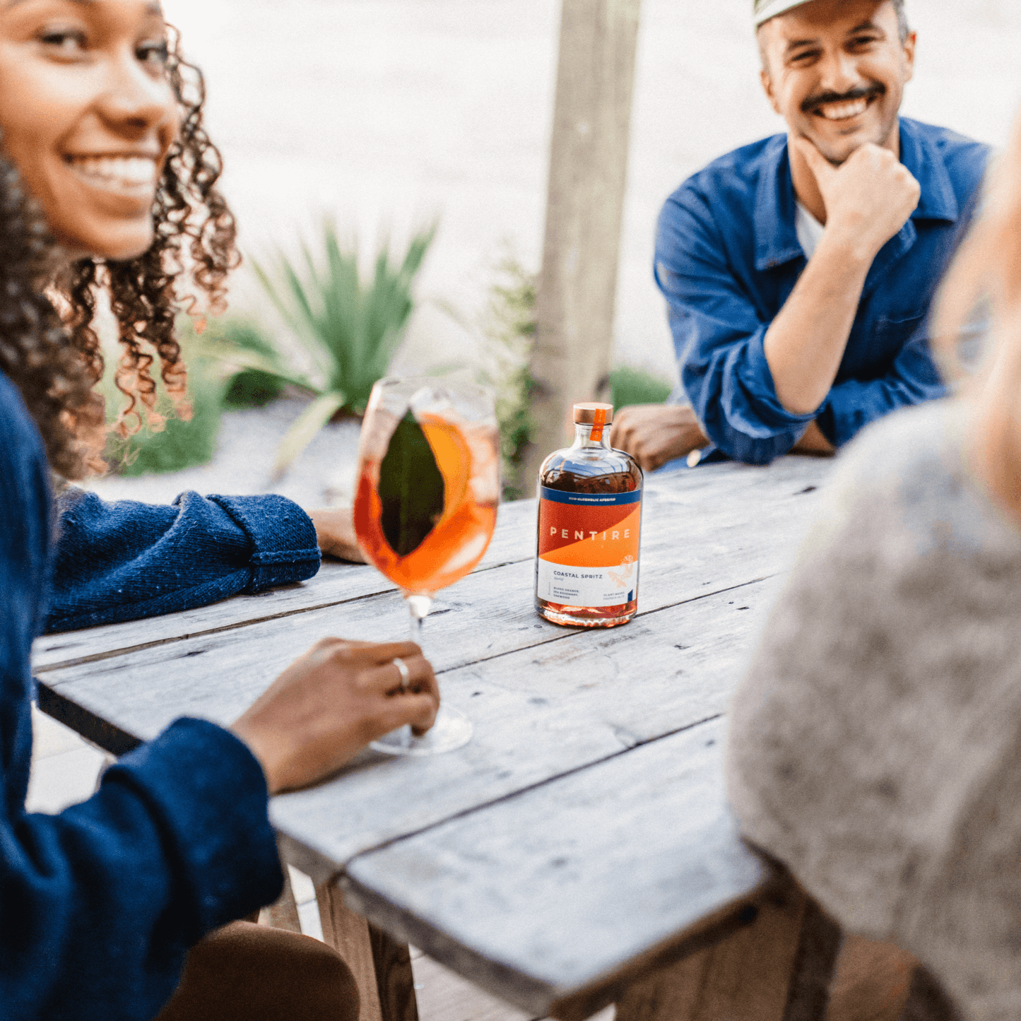 People enjoying Pentire Coastal Spritz, a refreshing non-alcoholic aperitif, at a rustic outdoor table.