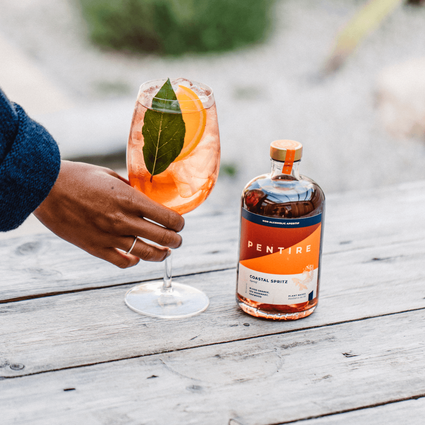 Person holding a glass of Pentire Coastal Spritz with a bottle on a wooden table outdoors.
