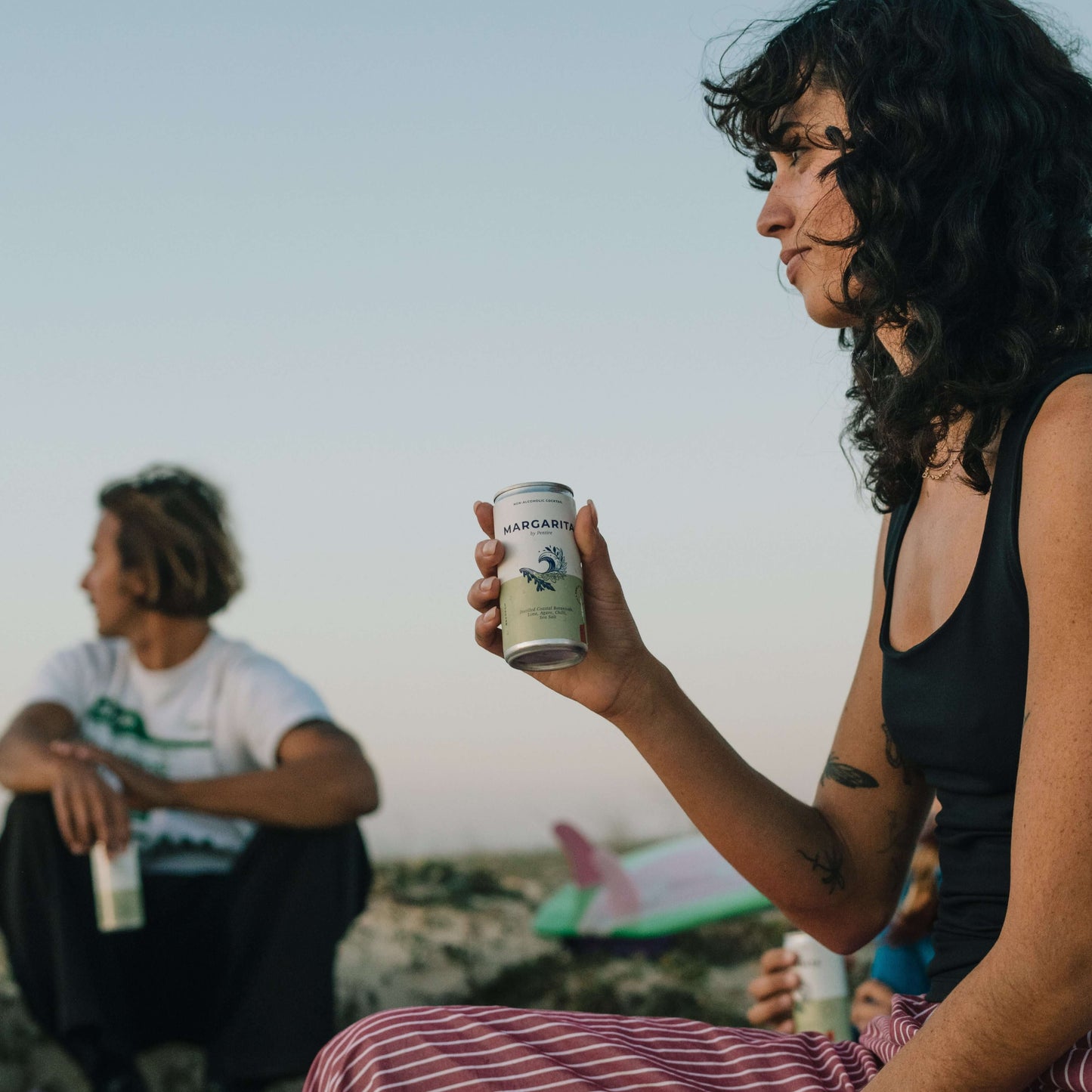 Person enjoying a Pentire Margarita can on a beach, with friends in the background, showcasing the refreshing cocktail.