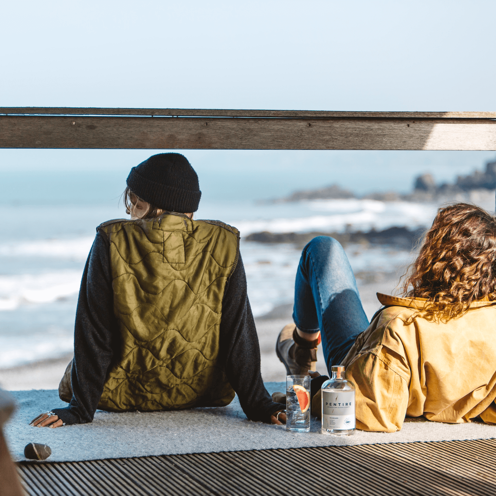 Two people enjoying Pentire Seaward non-alcoholic spirit by the ocean, dressed in cozy outdoor attire.
