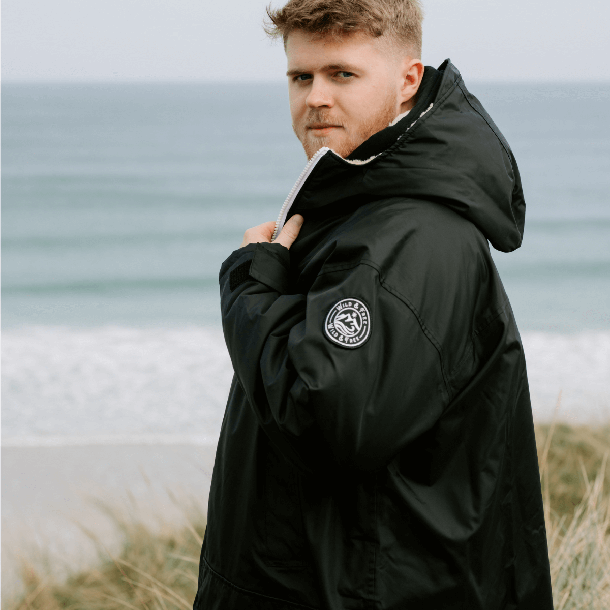 Raincoat & Changing Robe in One - The Classic Cobe Black model by the beach, showcasing its stylish design, eco-friendly materials.