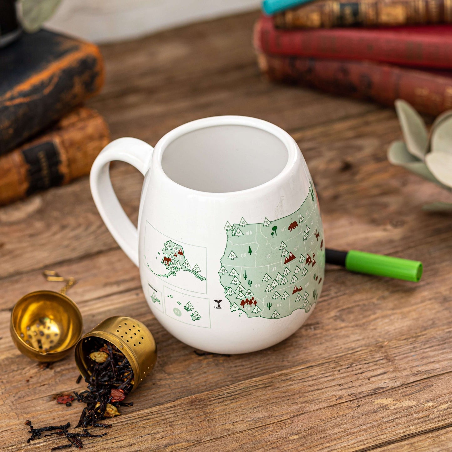 White mug with green illustrations on a wooden surface with books and tea accessories.