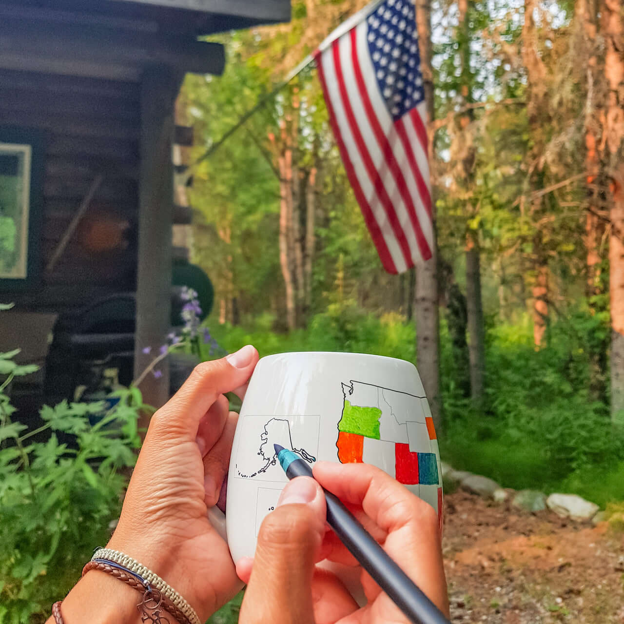 Person colouring in the USA Map Colour In Travel Mug with a black pen outdoors, surrounded by trees and an American flag.