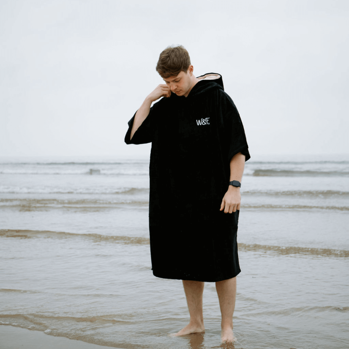 A person wearing The Luxury Towel Robe at the beach, enjoying the water in a stylish black beach towel robe.