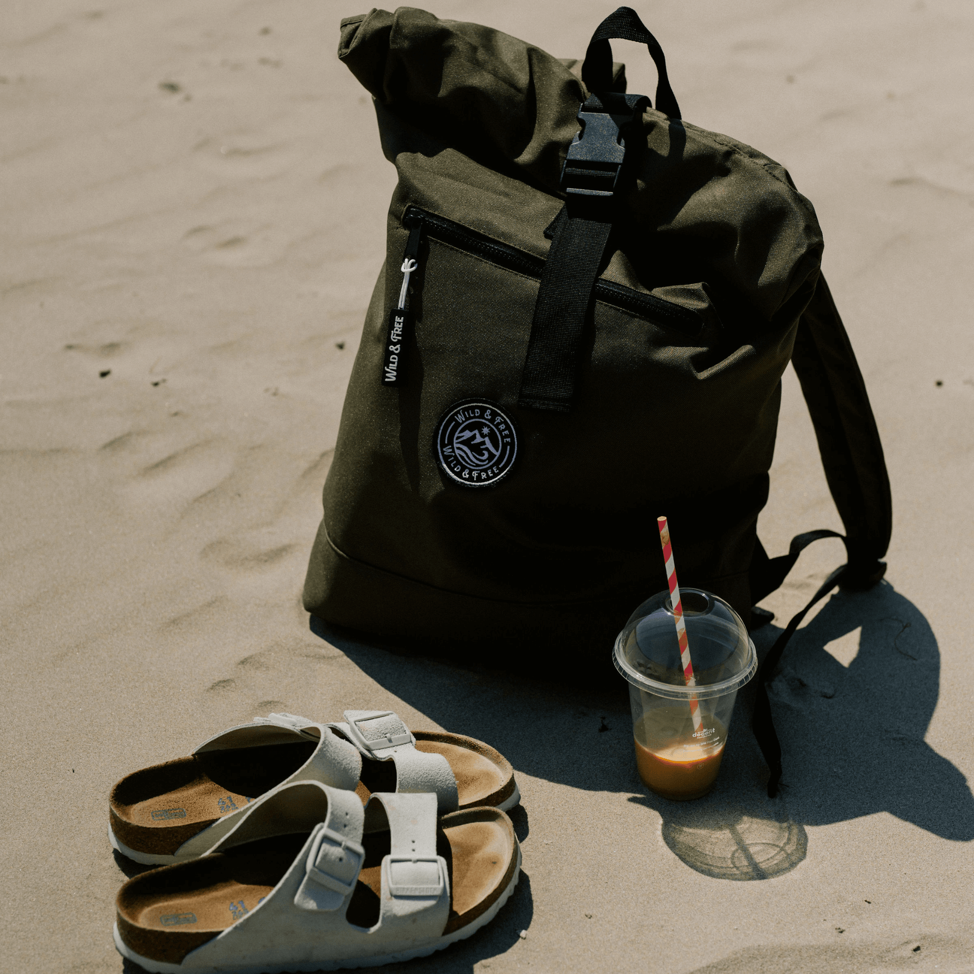 The Backpack by Wild & Free in an outdoor setting with sandals and a drink on the sand. Shop at Alewk.club.