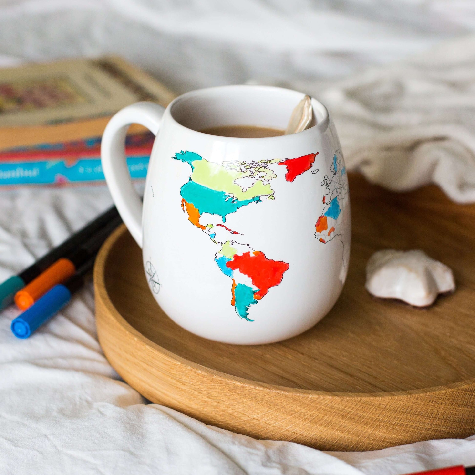White mug with world map design on a wooden tray with books and pens in the background.