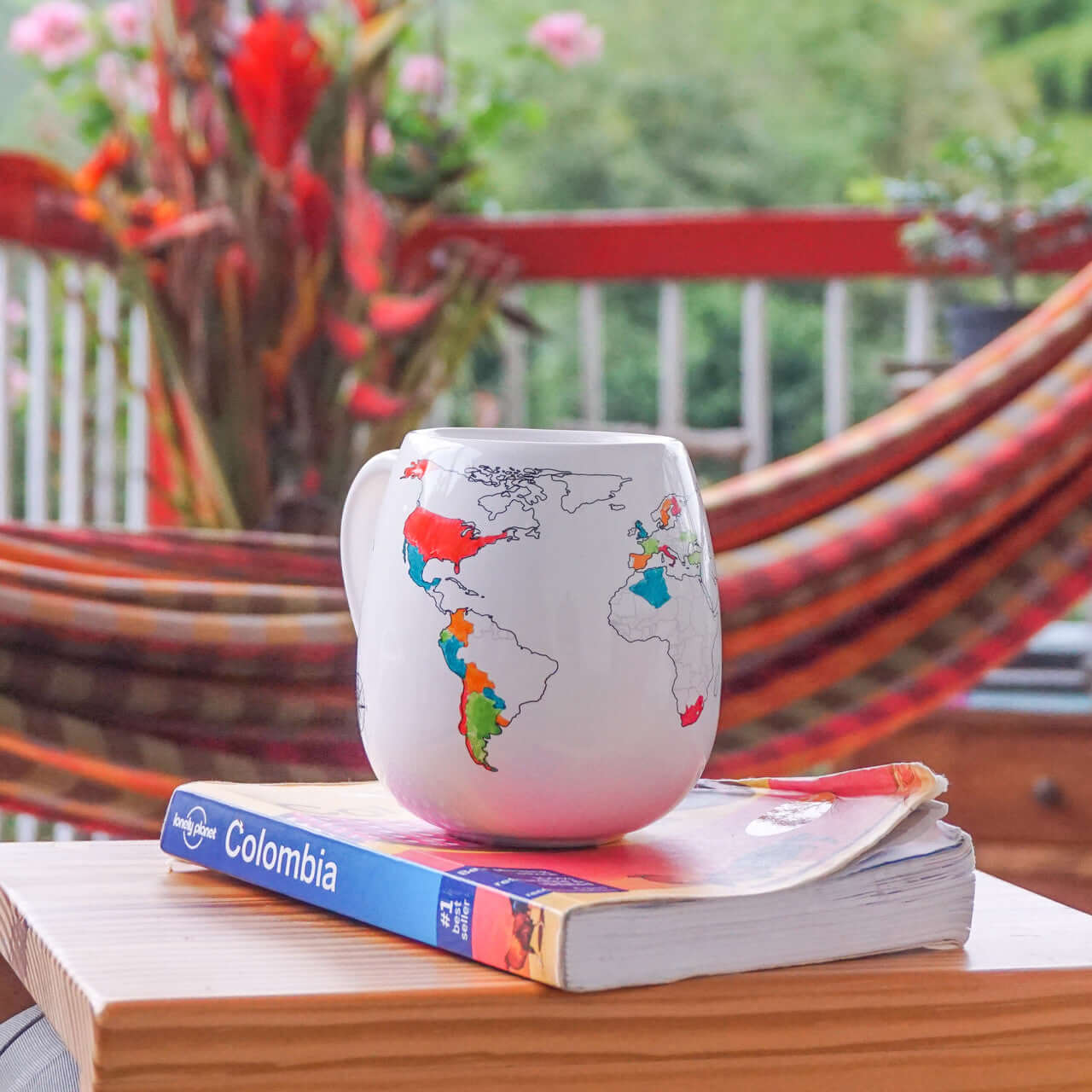 Stemless wine glass with world map design on a book with 'Colombia' on the cover, placed on a wooden surface with a blurred background of a hammock and plants.